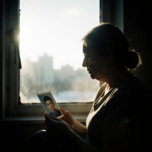 A woman gazes thoughtfully at a photo displayed on a window sill, surrounded by soft natural light.